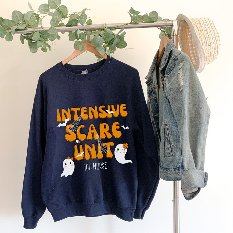 May include: Navy blue sweatshirt with the text "INTENSIVE SCARE UNIT" in orange and white, featuring bat, spiderweb, and ghost graphics. The text "ICU NURSE" is below the main text. A denim jacket and straw hat hang on the rack.