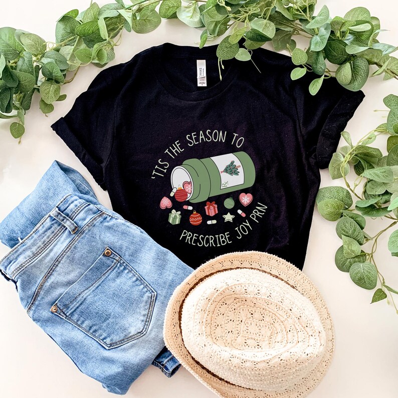 May include: Black t-shirt with the text "'Tis the Season to Prescribe Joy PRN" and a graphic of a medicine bottle spilling out Christmas-themed items. Light blue jeans and a straw hat are also in the image.