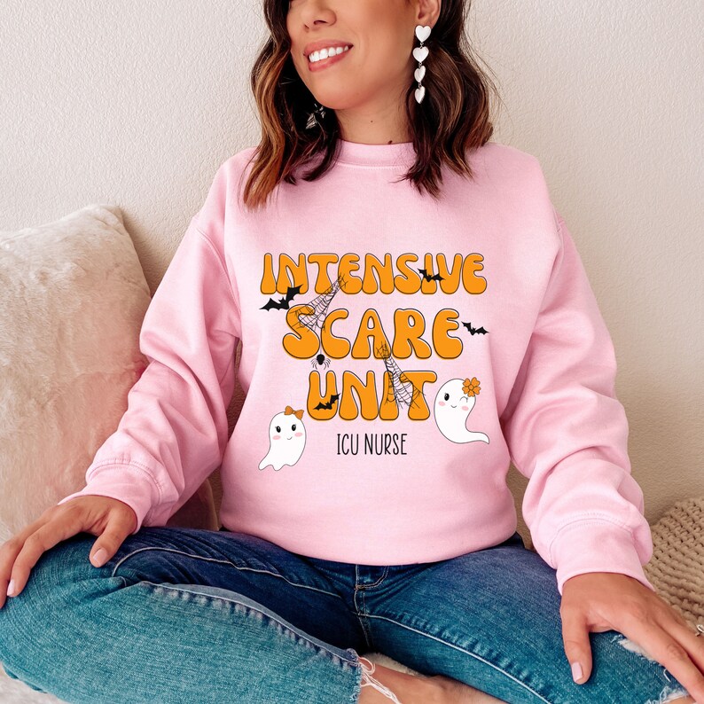 May include: A pale pink sweatshirt with the words "Intensive Scare Unit" in orange, with bats and spiderwebs. Two cartoon ghosts are also featured. The text "ICU Nurse" is below the main text. The sweatshirt is a Halloween-themed design.