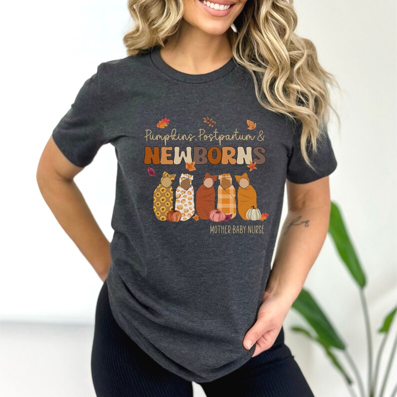 May include: Dark grey t-shirt with the text "Pumpkins, Postpartum & Newborns" in brown and orange lettering. The shirt features an illustration of five swaddled babies with pumpkins and autumn leaves. The words "Mother Baby Nurse" are below the illustration.