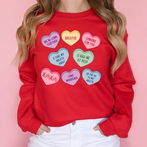 May include: A red sweatshirt with heart-shaped graphics in various colours. The hearts have phrases such as "Breathe" and "Lunging for You." The design is reminiscent of candy hearts, with lung-related puns.