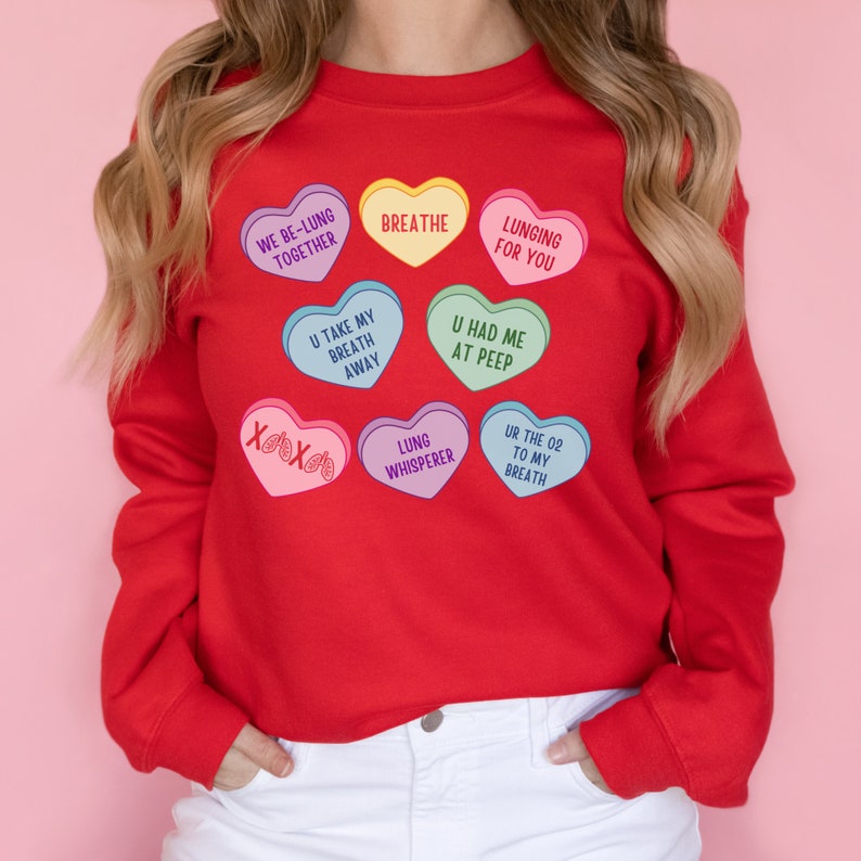 May include: A red sweatshirt with heart-shaped graphics in various colours. The hearts have phrases such as "Breathe" and "Lunging for You." The design is reminiscent of candy hearts, with lung-related puns.