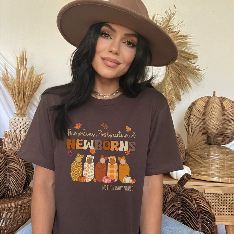 May include: A brown t-shirt featuring the words "Pumpkins, Postpartum & Newborns" in orange and brown. The design includes illustrations of swaddled babies and pumpkins. The text "Mother Baby Nurse" is at the bottom.