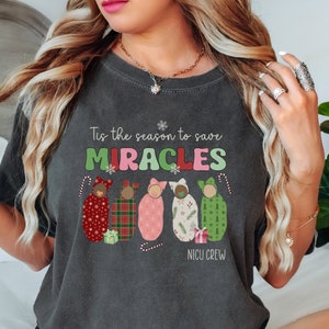 May include: Dark gray t-shirt with the text "Tis the season to save MIRACLES" in green, pink, and white. Below the text are five baby swaddles with holiday patterns and candy canes. The words "NICU CREW" are at the bottom.