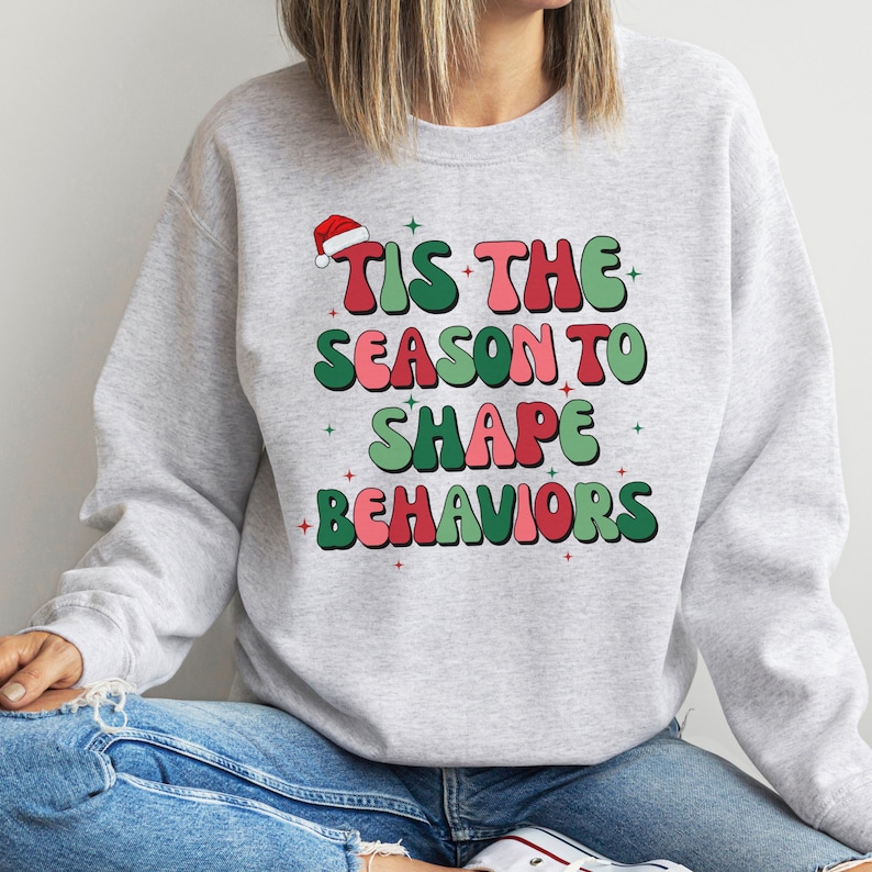 May include: A light gray sweatshirt with the text "TIS THE SEASON TO SHAPE BEHAVIORS" in red, green, and pink. A Santa hat is above the text. The sweatshirt is a casual, festive garment.