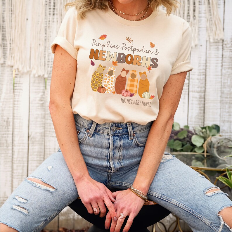 May include: A cream-coloured t-shirt featuring the text "Pumpkins, Postpartum & NEWBORNS" and an illustration of five swaddled babies. The design includes pumpkins and autumn leaves. The words "Mother Baby Nurse" are also printed on the shirt.