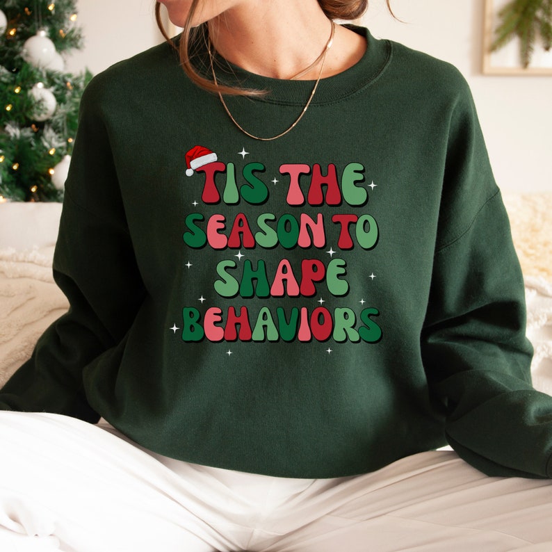 May include: Dark green sweatshirt with the text "TIS THE SEASON TO SHAPE BEHAVIORS" in red, green, and pink, with a Santa hat on top. The sweatshirt is a festive holiday design.