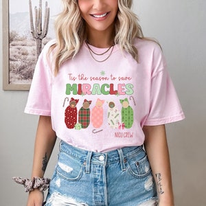May include: A light pink t-shirt with the text "Tis the season to save MIRACLES" in green and red. Below the text are five baby swaddles in various patterns and colors, with the words "NICU CREW" below them.