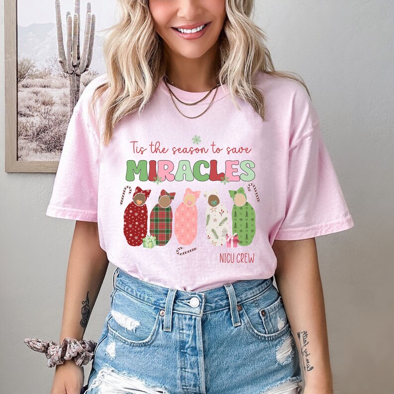 May include: A light pink t-shirt with the text "Tis the season to save MIRACLES" in green and red. Below the text are five baby swaddles in various patterns and colors, with the words "NICU CREW" below them.