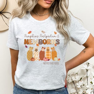 May include: Pale grey t-shirt featuring the text "Pumpkins, Postpartum & NEWBORNS" in brown and orange. The design includes illustrations of newborns wrapped in blankets, with pumpkins and autumn leaves.