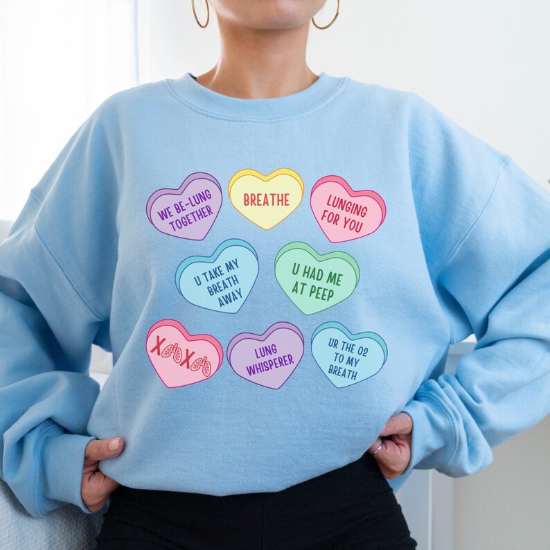 May include: Pale blue sweatshirt featuring heart-shaped graphics with phrases like "WE BE-LUNG TOGETHER" and "BREATHE." The design includes various colours and playful text. A relaxed fit, comfortable garment.