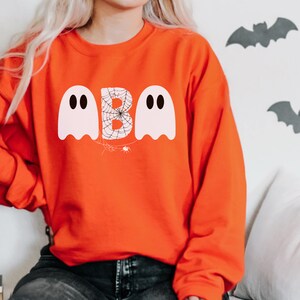 May include: Orange sweatshirt with a Halloween-themed design. The design features the word "BOO" with the letter "B" designed as a spider web, and two white ghost figures. The sweatshirt is a crew neck style.