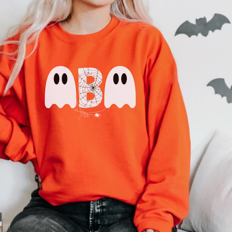 May include: Orange sweatshirt with a Halloween-themed design. The design features the word "BOO" with the letter "B" designed as a spider web, and two white ghost figures. The sweatshirt is a crew neck style.