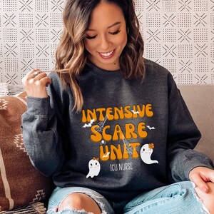 May include: Dark grey sweatshirt featuring the words "INTENSIVE SCARE UNIT" in orange and white, with bat, spiderweb, and ghost graphics. The text "ICU NURSE" is below. The design is a Halloween theme.