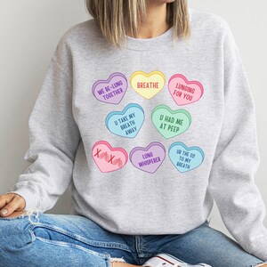 May include: A pale grey sweatshirt with a design of colourful heart-shaped candies. The hearts feature phrases such as "Breathe" and "Lunging for You." The design includes hearts in various colours, such as yellow, pink, and blue, with playful text.