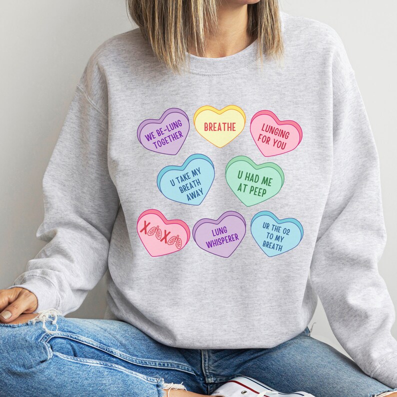 May include: A pale grey sweatshirt with a design of colourful heart-shaped candies. The hearts feature phrases such as "Breathe" and "Lunging for You." The design includes hearts in various colours, such as yellow, pink, and blue, with playful text.