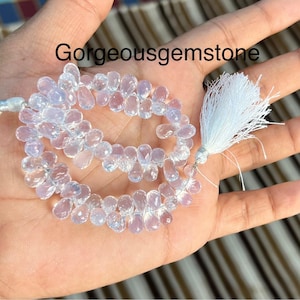 Könnte beinhalten: Eine Strähne aus klaren, facettierten, tropfenförmigen Perlen mit einem weißen Quasten an jedem Ende. Die Perlen sind auf einem silbernen Faden aufgefädelt. Gorgeousgemstone ist in schwarzer Schrift auf dem Bild zu sehen.