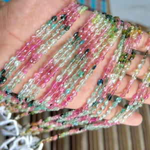 Könnte beinhalten: Stränge farbiger Edelsteinperlen in Rosa, Grün, Blau und transparent. Die kleinen, ovalen Perlen sind zu Halsketten oder Armbändern aufgereiht. Die Edelsteine werden in einer Hand gehalten.