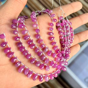 Puede incluir: Hebras de vibrantes cuentas de piedras preciosas rosas, cada una cortada en forma de lágrima. Las cuentas están ensartadas en un fino alambre plateado, creando una apariencia delicada y elegante. Las piedras preciosas se exhiben sobre un fondo neutro.