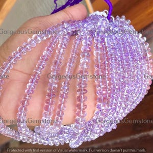 Könnte beinhalten: Stränge aus hellvioletten Edelsteinperlen. Die kleinen, runden, facettierten Perlen sind zu mehreren Strängen aufgereiht. Die Perlen werden in einer Hand gehalten, mit dem Text "Gorgeous Gemstone" sichtbar.