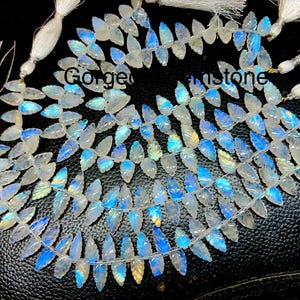 May include: A strand of white gemstone beads with a leaf shape and iridescent blue and gold hues. The beads are strung on a clear thread and have a tassel at each end.