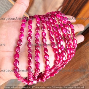 Puede incluir: Una colección de vibrantes cuentas de piedras preciosas de rubí de forma ovalada. Las cuentas son de un color rojo-rosado intenso y rico y están ensartadas, creando un collar o un hilo de piedras preciosas. La imagen muestra la belleza natural y el color de los rubíes.