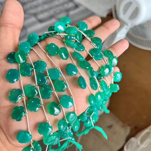 Pode incluir: Um fio de contas de pedras preciosas verdes, cada conta tem o formato de uma flor. As contas são enfiadas em um cordão branco com detalhes dourados.