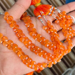 May include: Close-up of several strands of orange gemstone beads. The beads are teardrop-shaped and strung together. The strands are held in a hand, with orange tassels visible at the top. The background is a striped fabric.