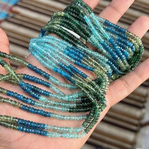 以下が含まれることがあります： 緑と青の色合いの宝石ビーズのストランドが表示されています。 ビーズは小さくて丸く、ネックレスを形成するために一緒にひもでつながれています。 色のグラデーションは、濃いティールから明るいターコイズまであります。