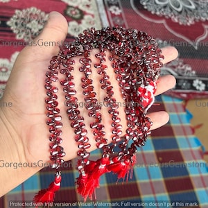 Puede incluir: Un collar de cuentas de piedras preciosas de color rojo intenso con una borla. Las cuentas son pequeñas y de forma ovalada, ensartadas para formar múltiples hebras. Las borlas son de un color rojo vibrante. La imagen es de una mano sosteniendo las cuentas.