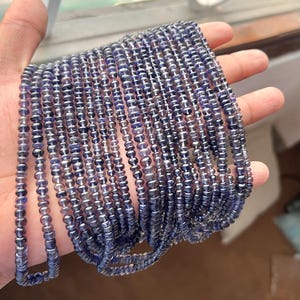 Könnte beinhalten: Eine Strähne aus blauen und violetten Edelsteinperlen. Die Perlen sind klein und rund und auf einem durchsichtigen Faden aufgereiht. Die Perlen sind in einem lockeren, fließenden Muster angeordnet.