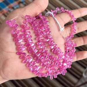 Könnte beinhalten: Stränge aus rosa Edelsteinperlen, jede tropfenförmig. Die Perlen sind zu einer Halskette oder einem Armband aufgereiht. Die Edelsteine haben eine leuchtend rosa Farbe, und die Perlen sind glatt und poliert.