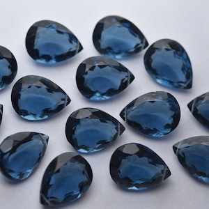 Könnte beinhalten: Eine Sammlung von 15 tropfenförmigen blauen Edelsteinen. Die Edelsteine sind facettiert und haben einen perlmuttartigen Glanz.