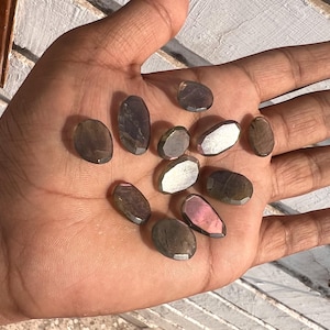 Peut inclure: Une collection de 10 pierres précieuses de labradorite de forme ovale dans différentes nuances de gris, noir et rose. Les pierres sont lisses et polies, avec un léger éclat iridescent.