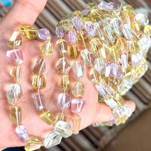 May include: A multi-strand necklace with faceted, irregular beads in yellow, clear, and lavender. The beads are strung on gold wire. The necklace is held in a hand. The necklace is a piece of jewellery.