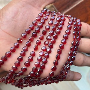 Pode incluir: Várias fileiras de contas de pedras preciosas facetadas, vermelho escuro, são seguradas em uma mão. As contas em forma de gota são enfiadas, para um colar ou pulseira. As pedras têm uma qualidade rica e translúcida.