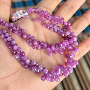May include: A close-up of a necklace made of small, faceted, purple beads. The beads are strung together in a teardrop shape. The necklace is held in a hand, with the background out of focus.