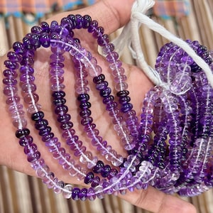 Könnte beinhalten: Stränge aus Amethystperlen in verschiedenen Lila- und Klartönen. Die kleinen, runden Perlen sind auf weißem Faden aufgereiht. Die Perlen werden in einer Hand gehalten, der Hintergrund ist unscharf.