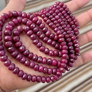May include: Strands of faceted, deep red ruby beads. The beads are strung together, forming multiple rows of the gemstone. The ruby beads are a rich, saturated color, and the facets catch the light, creating a sparkling effect. The beads are held in a hand.