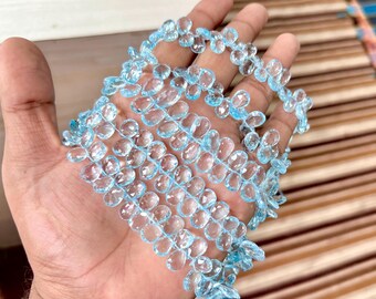 May include: A strand of light blue, teardrop-shaped gemstones, possibly topaz, is held in a hand. The gems are strung together, creating a delicate, sparkling necklace or bracelet. The gems are approximately 0.5 inches in length.
