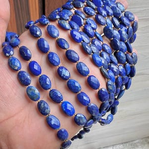 Puede incluir: Una colección de cuentas de lapislázuli facetadas, de forma ovalada y en varios tonos de azul. Las cuentas están ensartadas con espaciadores dorados, creando un collar o un hilo de piedras preciosas. Las piedras preciosas se sostienen en una mano.