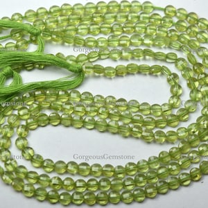 Könnte beinhalten: Eine Strähne grüner Peridot-Perlen, etwa 6 mm groß, mit facettierten Oberflächen. Die Perlen sind auf einer dünnen Schnur aufgefädelt und haben an jedem Ende einen Quasten. Die Perlen liegen lose auf einer weißen Oberfläche.