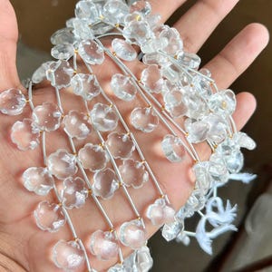 Puede incluir: Una hebra de cuentas de cuarzo claro en forma de flor enhebradas en un alambre plateado. Las cuentas son facetadas y tienen una apariencia translúcida.