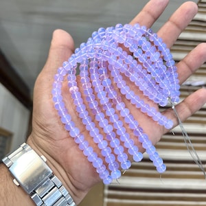 Könnte beinhalten: Stränge hellvioletter, durchscheinender Perlen. Die runden Perlen sind aufgefädelt. Die Perlen werden in einer Hand gehalten, wobei eine silberne Armbanduhr am Handgelenk zu sehen ist.