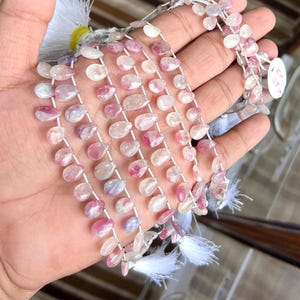Könnte beinhalten: Stränge aus rosa und weißen, tropfenförmigen Edelsteinen, auf weißem Faden aufgereiht. Die Edelsteine werden in einer Hand gehalten, wodurch ihre zarten Farbvariationen zur Geltung kommen. Die Perlen sind wahrscheinlich für die Schmuckherstellung bestimmt.