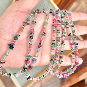 Könnte beinhalten: Eine Sammlung farbenfroher Turmalin-Halsketten. Die Halsketten bestehen aus kleinen, unregelmäßig geformten Steinen in Rosa-, Grün-, Blau- und Schwarztönen. Die Halsketten werden auf einer Hand präsentiert.