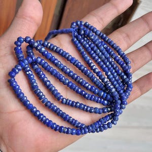 Puede incluir: Una colección de cuentas facetadas de lapislázuli azul intenso. Las cuentas están ensartadas, formando múltiples hebras. Las cuentas de piedras preciosas se sostienen en una mano, mostrando su color y textura. Las cuentas son de un azul intenso.