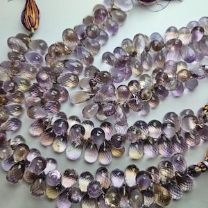 Könnte beinhalten: Eine Strähne aus Amethyst-Edelsteinkugeln in Tropfenform. Die Perlen sind ein Mix aus hell- und dunkelvioletten Farbtönen mit einem leichten Braunton. Die Perlen sind auf einer Schnur mit Quasten an jedem Ende aufgefädelt.