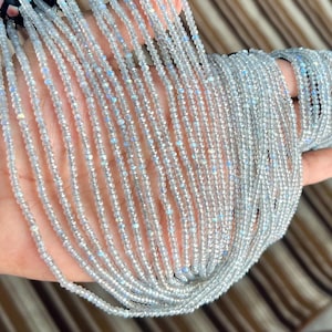 Könnte beinhalten: Eine Sammlung zarter, hellgrauer Edelsteinperlen. Die Perlen sind in mehreren Strängen aufgereiht, wodurch ein schimmernder Effekt entsteht. Die Perlen werden in einer Hand gehalten, um ihre Farbe und Textur zu zeigen. Das Gesamtthema ist Schmuck.