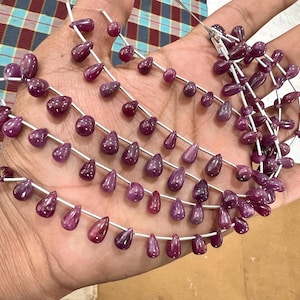 Op de afbeelding: Een streng paarse edelsteen kralen in een druppelvorm. De kralen zijn geregen op zilverdraad en zijn gerangschikt in een los, vloeiend patroon.